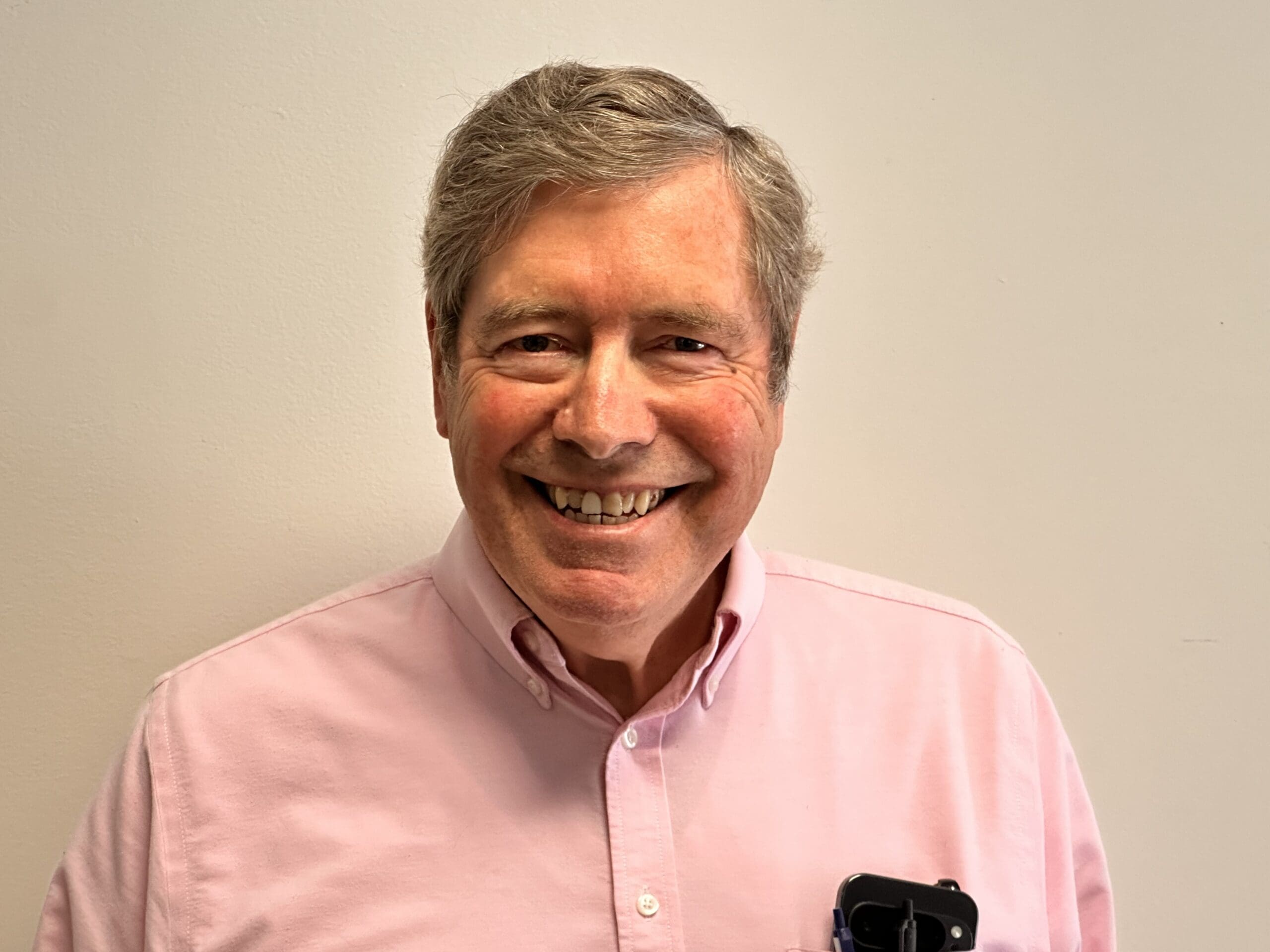 Smiling middle-aged man with gray hair wearing a pink button-down shirt, standing against a beige wall; a phone is visible in his chest pocket.