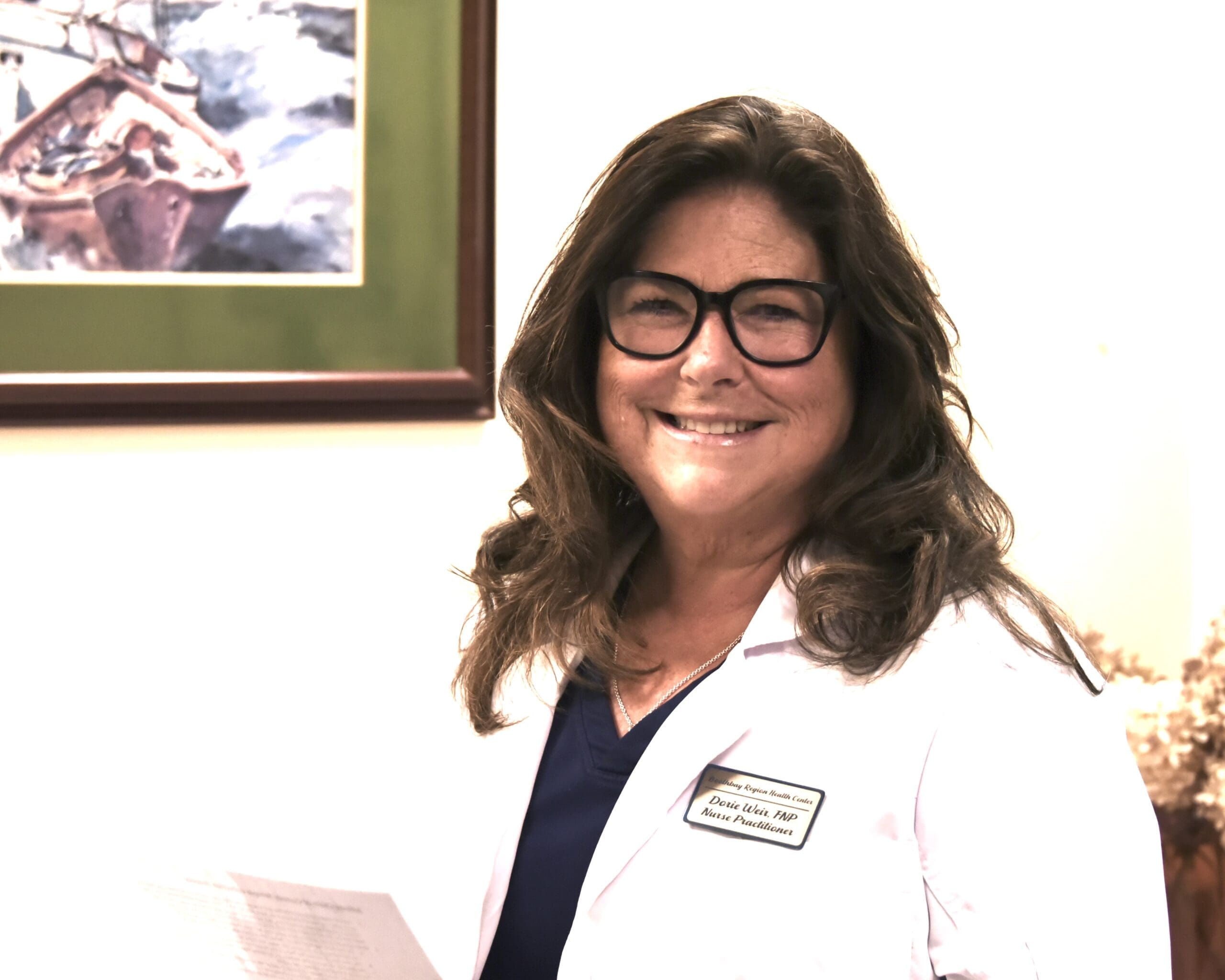 Smiling woman wearing glasses and a white medical coat with a name badge visible on her chest.